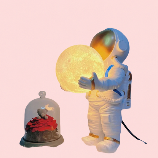 Astronaut holding a moon-shaped lamp with a small decorative scene below on a pink background