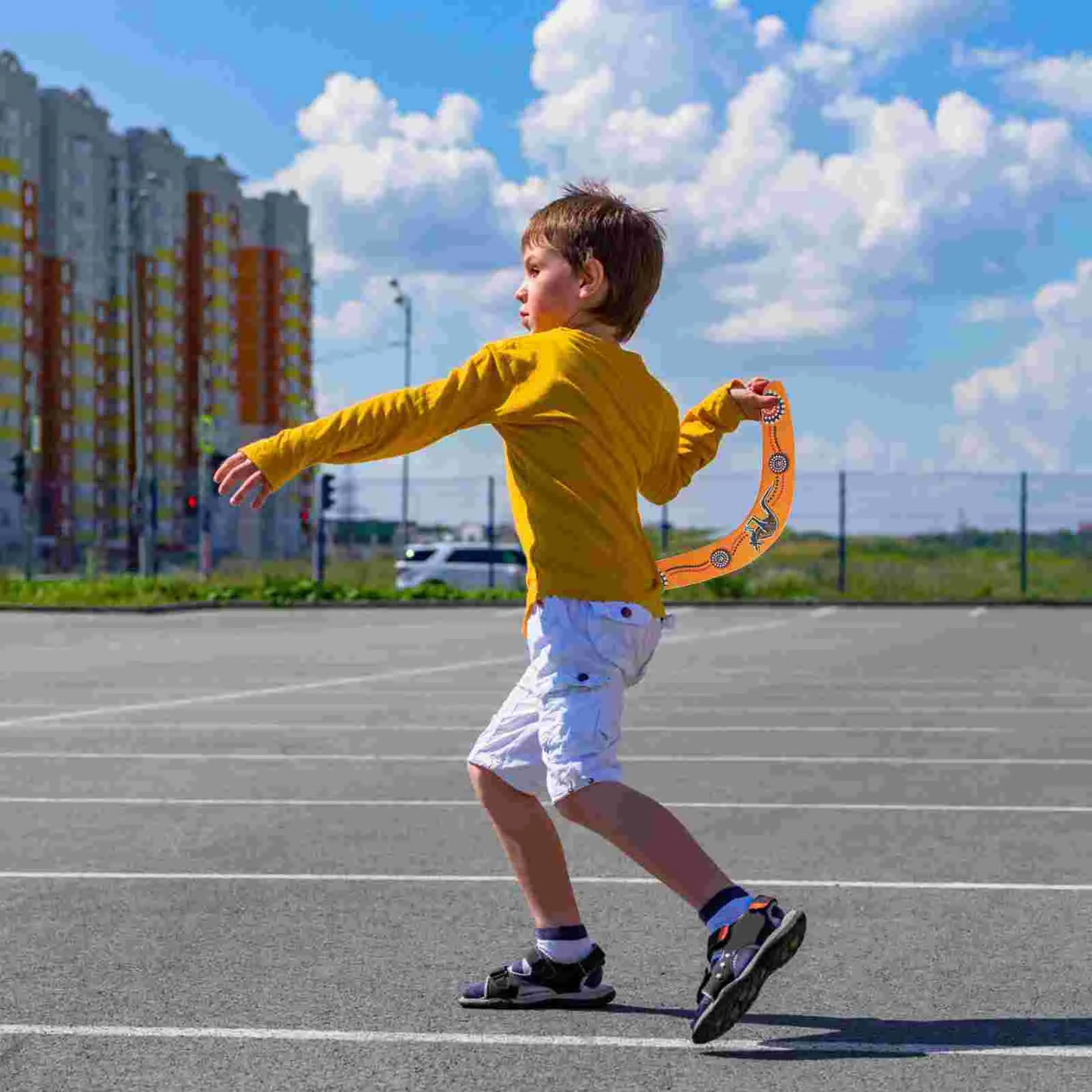Child in a yellow shirt and white shorts running on a paved area with buildings and clouds in the background to thrrough Decorative boomerang with Aboriginal art design held by a hand.